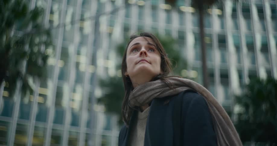 A woman wearing a scarf and a coat standing outdoors with a background of tall buildings and palm trees