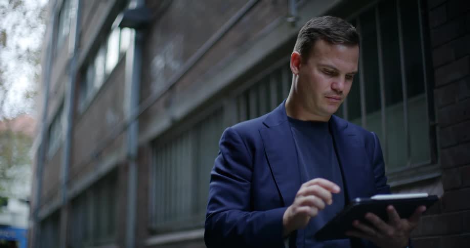 A man in a blazer uses a tablet outside a city building