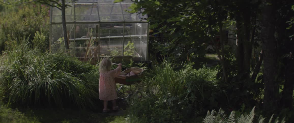A young girl pushes a baby carriage across a yard and garden