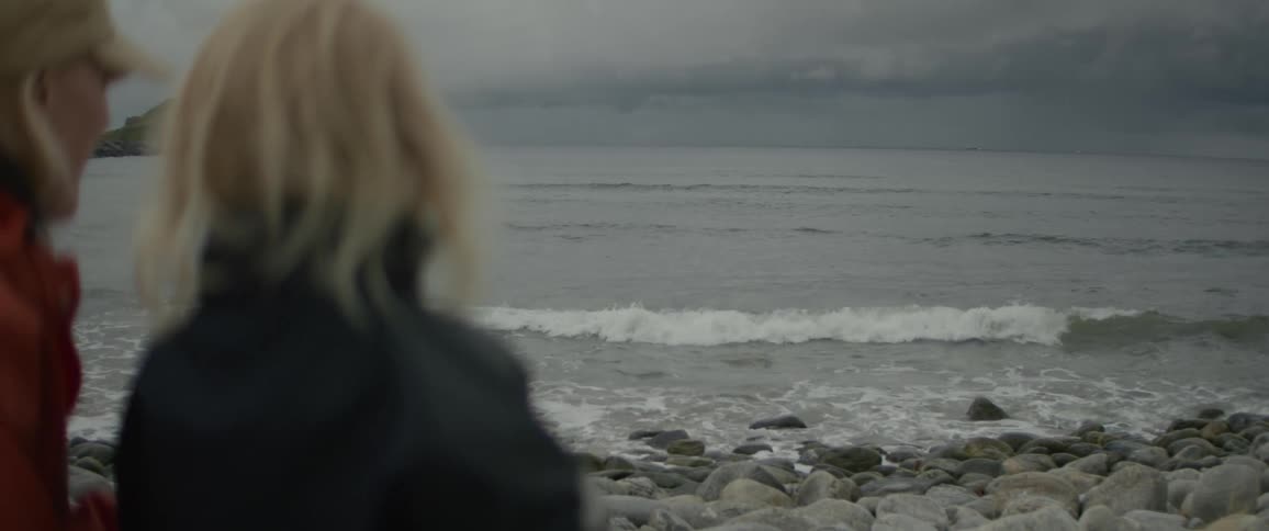 A person with blonde hair stands on a rocky beach facing the ocean with waves crashing against the shore on a cloudy day