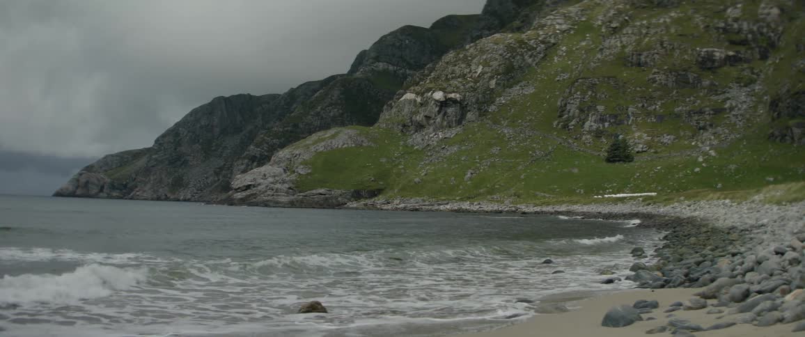 Waves foam while rolling onto a beach partially covered in stones near seaside mountains on an overcast day
