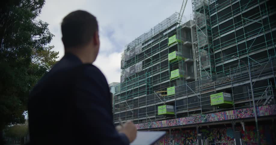 A man in a suit uses a stylus to take notes on a tablet in front of a building under construction