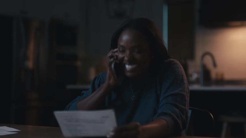 A woman talks on a phone laughing and smiling about something on a piece of paper