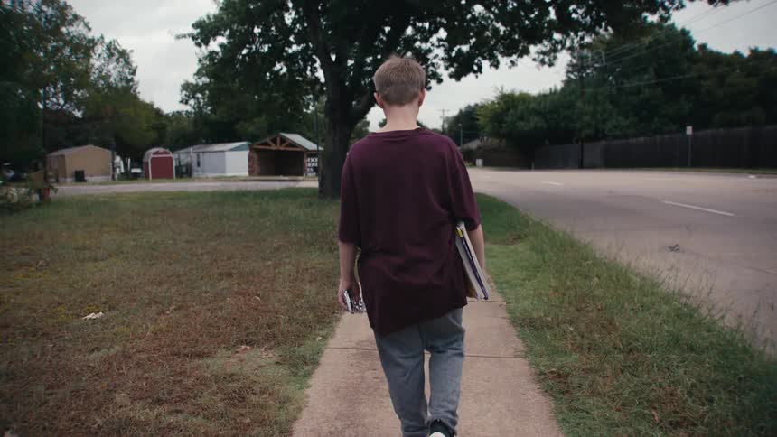 Back view of a boy walking on the sidewalk of a residential neighborhood holding a briefcase
