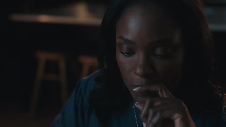 A woman in a blue shirt and wearing a necklace is looking forward and she is in an indoor environment that is dark behind her