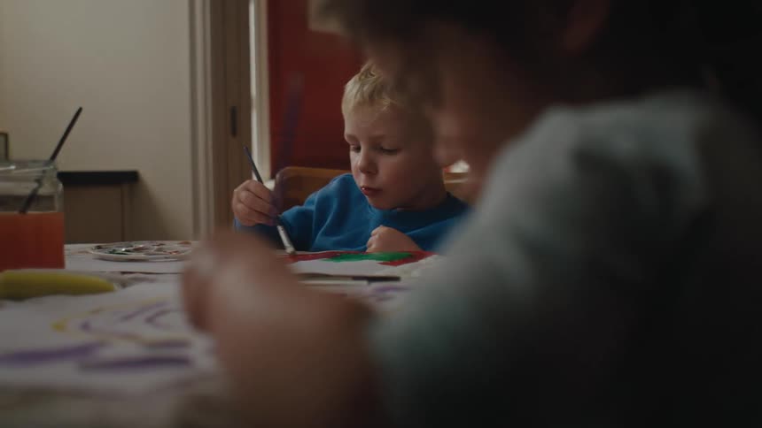 Two little children sitting at the table holding a paintbrush and painting with paint