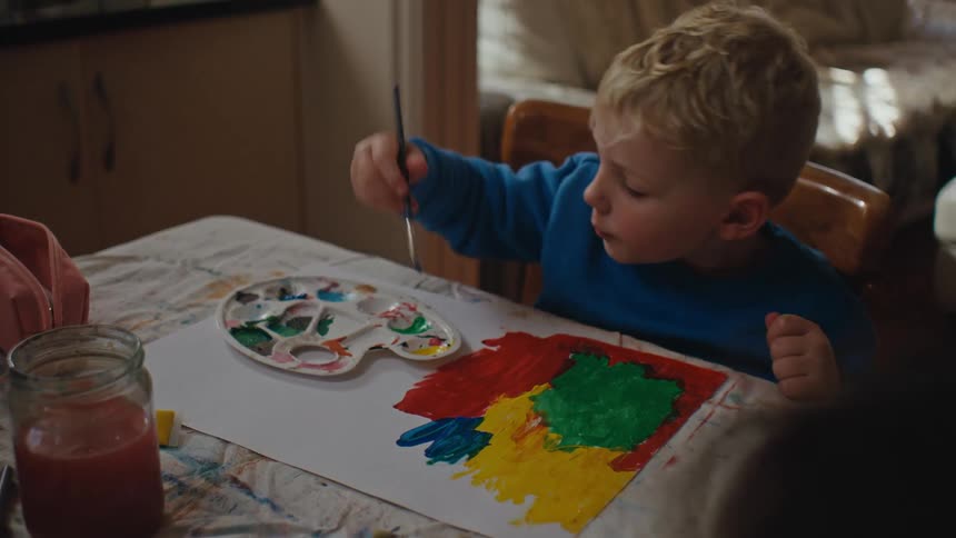 Little blond boy sitting at the table painting with paint and a brush