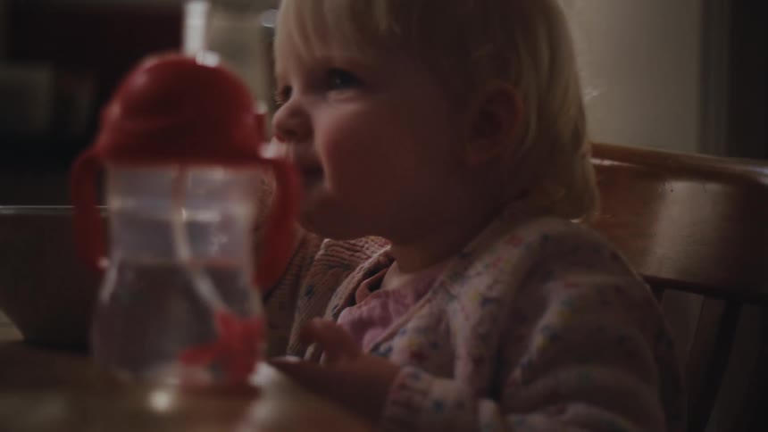 A little blonde girl is chewing something while sitting looking forward and on the table in front of her there is a bottle