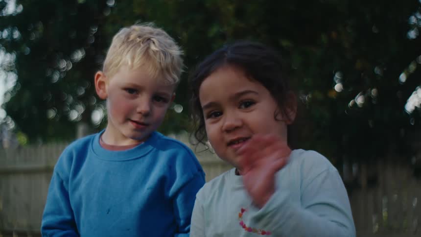 A little blond boy and a little brunette girl are looking forward in an outdoor setting and the little girl is signing and they have a little smile