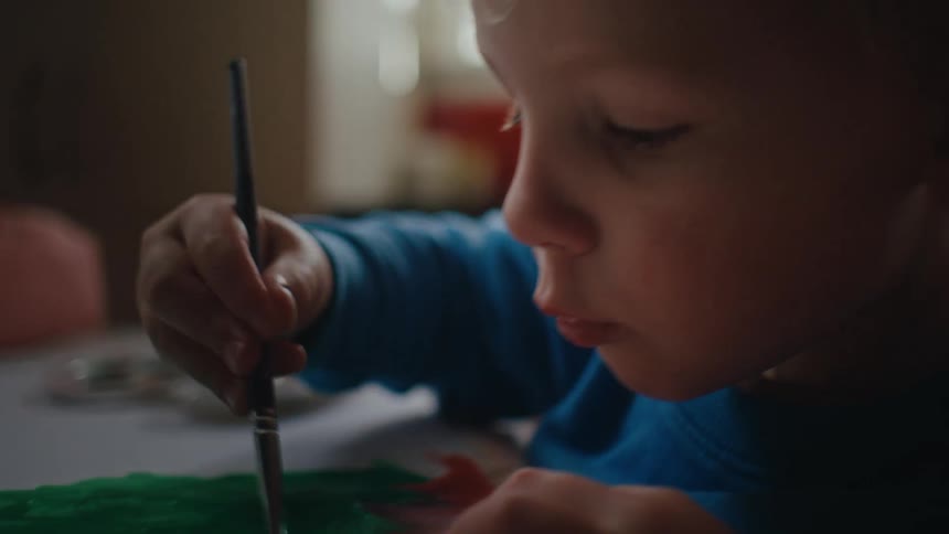 The little blond boy in the blue shirt is painting a paper with green and brown paint using a brush