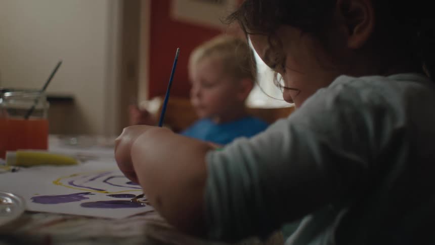 Two children sitting at a table drawing or painting on paper