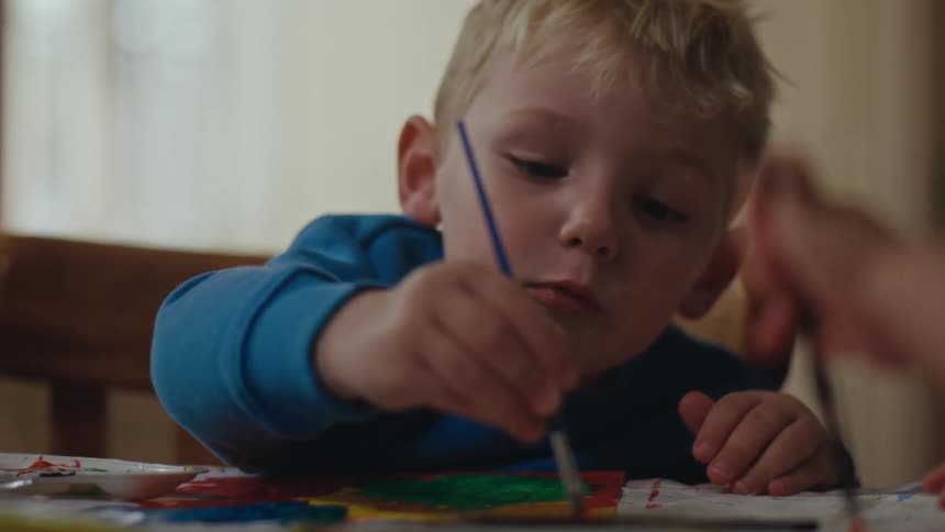 Little blond boy with a brush painting on a white sheet on the table
