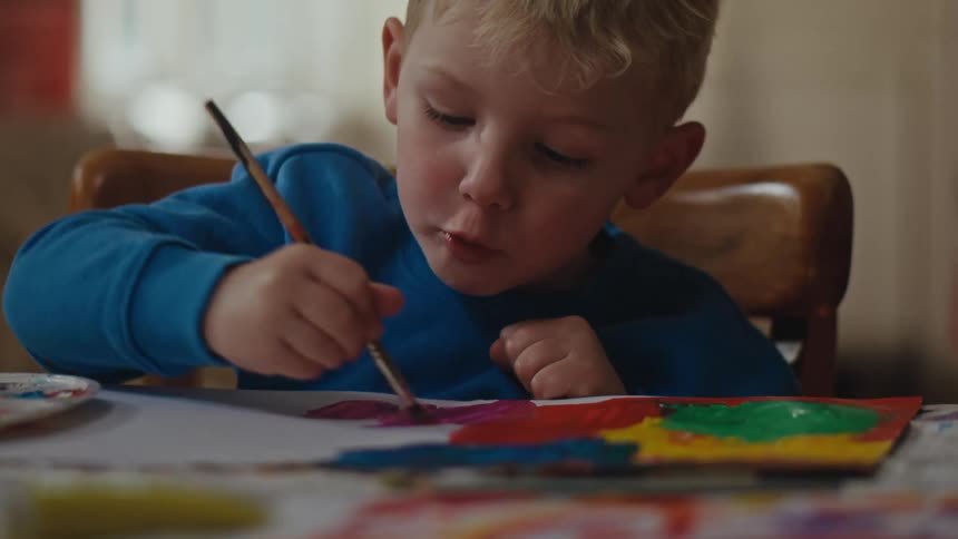 Boy sitting at table holding a paintbrush and painting a blank sheet of paper