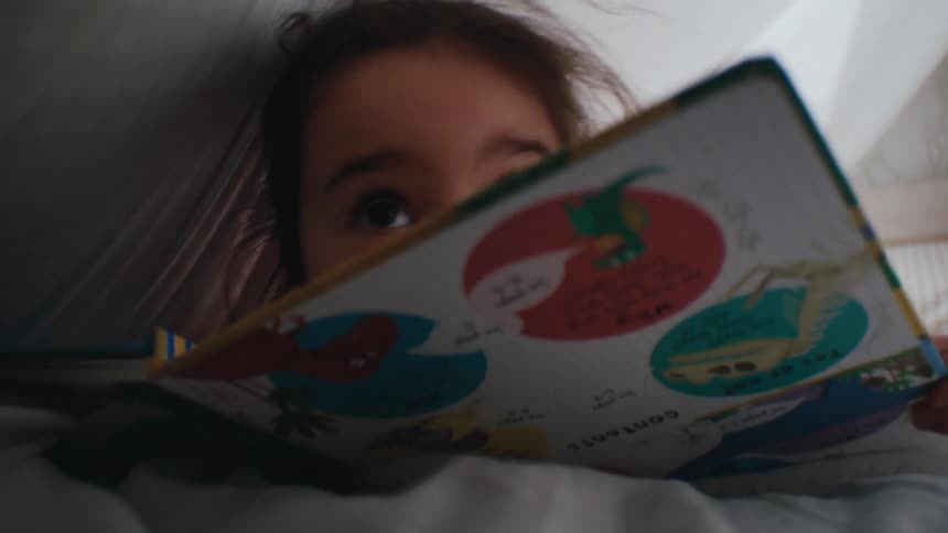 A little girl is under a white blanket smiling while covering her face with a childrens book