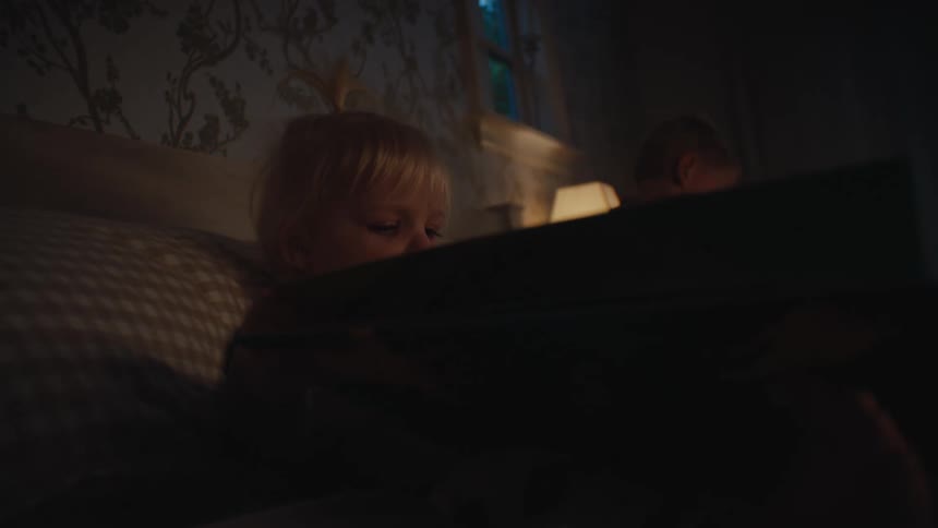Two children one blonde and the other dark haired laying closely in a bed while reading books in a dimly lit room with patterned wallpaper