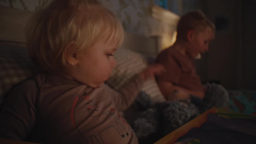 Two little blond boys lying in a bed with blankets and the smaller boy is snuggling in the bed