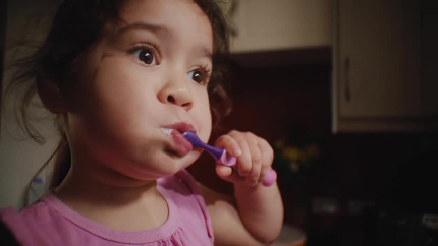 A little girl staring straight ahead while brushing her teeth using a purple and pink toothbrush