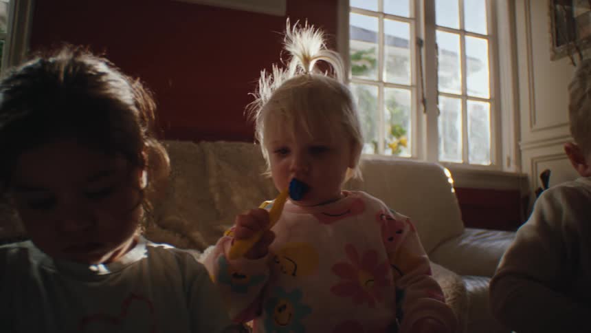 Blonde girl sucks on a toy while two other children concentrate on their activities as they all sit in the living room