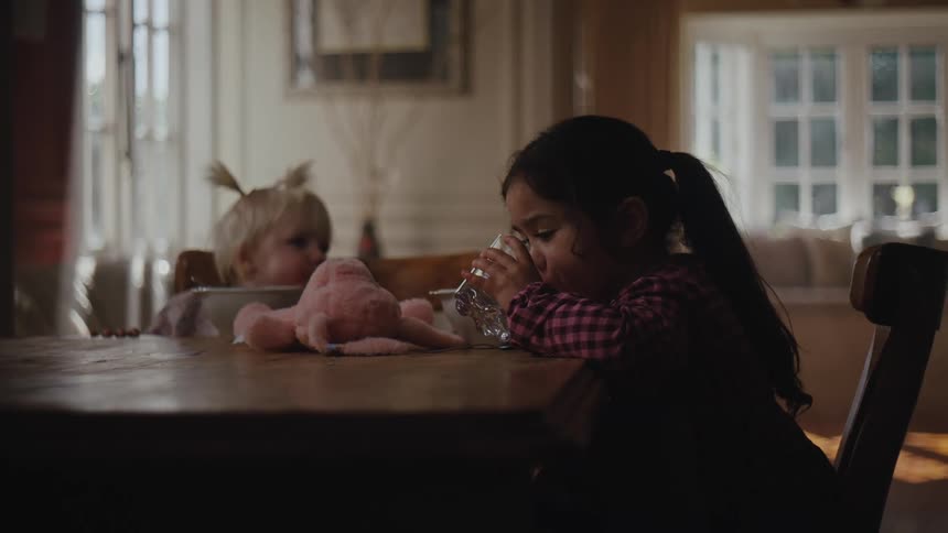 Two small children sitting at a table with one of them drinking water
