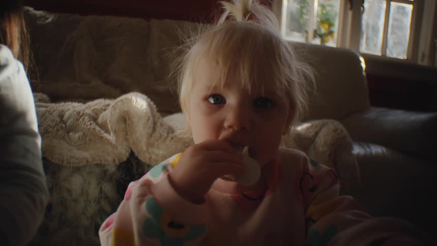 A female toddler sits next to an older female while chewing on a pacifier in a living room