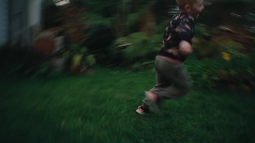 A little blonde boy is running on the grass in a backyard