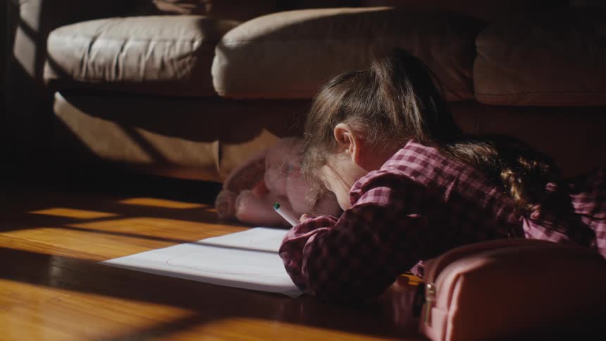 A little girl lying on a wooden floor in the living room drawing on paper