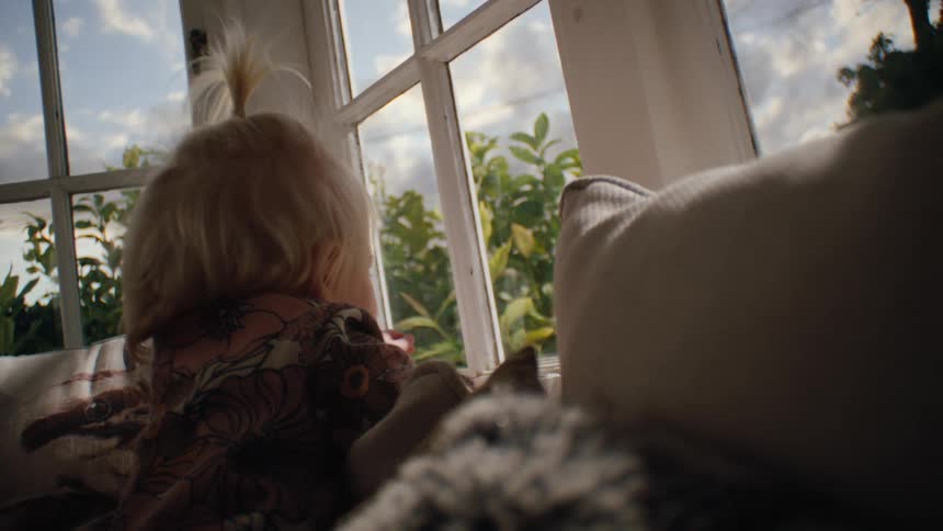 Little blonde girl on the couch among the cushions next to a window