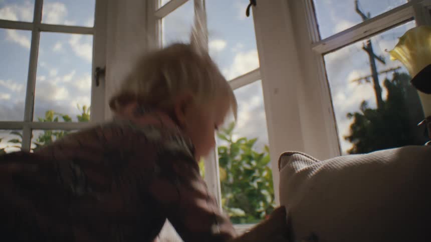 A child climbs and jumps over a couch near a window in a house