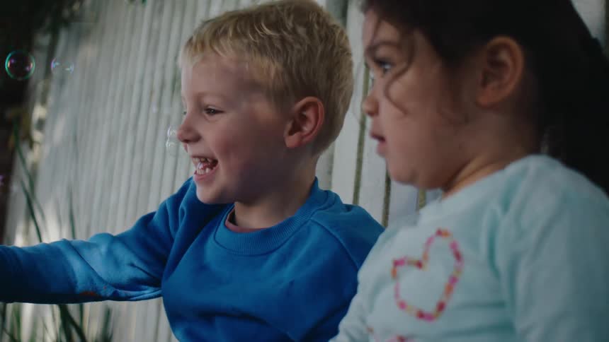 A couple of children recline on a white fence in the backyard playing with soap bubbles