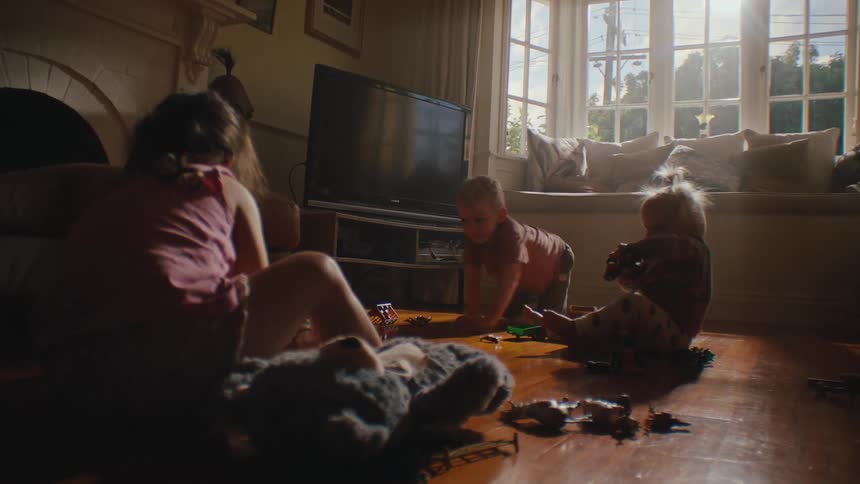 Three young siblings play together on the wooden floor of a living room