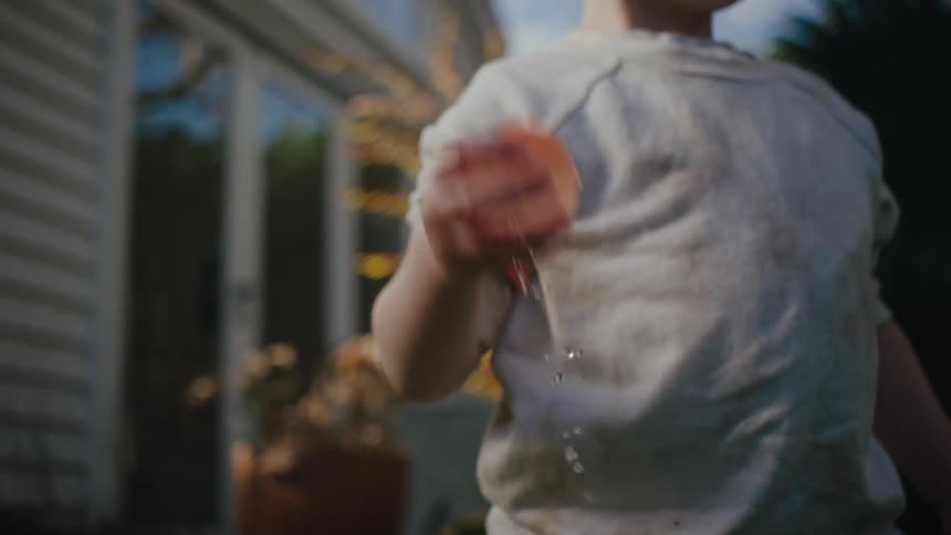 A little blond boy wearing a white coat is picking up a balloon filled with water from a bucket and throwing it forward