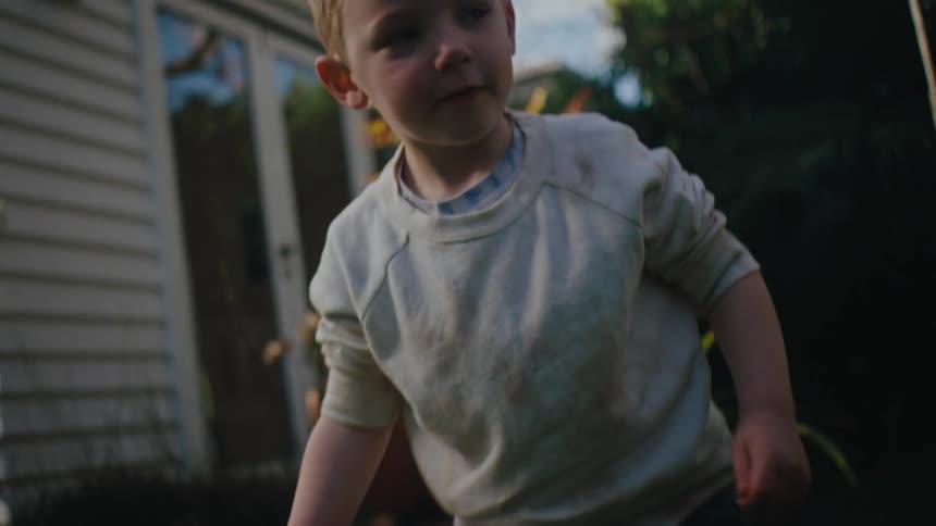 A little blond boy is in a backyard messing with a metal bucket and then he gets up