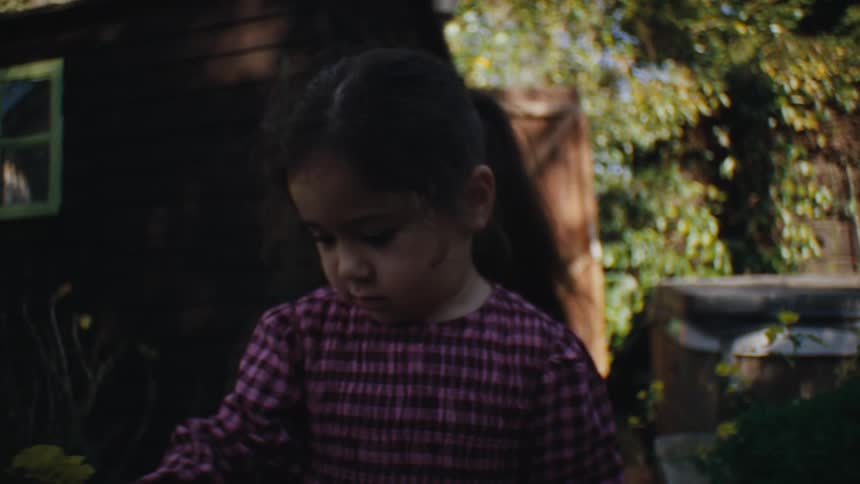 A little girl in checkered clothes is picking up a plant with yellow flowers and pulling it