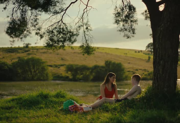 A man and a woman are sitting under a tree on the grass next to a river and talking and the man is shirtless