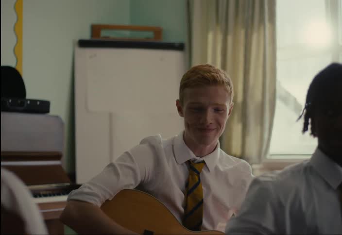 A young blond man in a white shirt and yellow tie holding a guitar is looking forward and has a smile