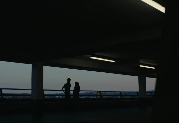 Two silhouetted figures standing in a parking garage with a large open window revealing a distant cityscape at dusk