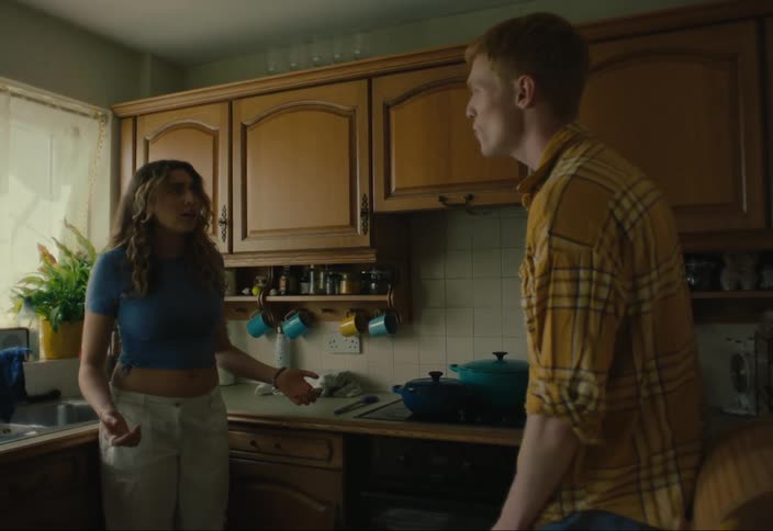 A man wearing a brown shirt argues with a woman with wavy hair and a blue tshirt in the kitchen