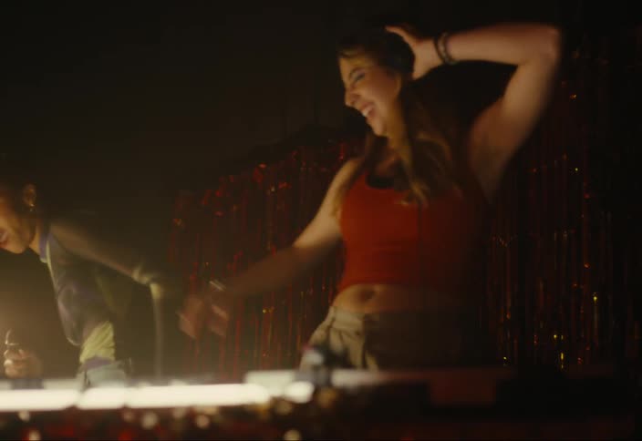 Two female djs dance behind a setup with turntables and a mixer set against a backdrop of shiny metallic streamers