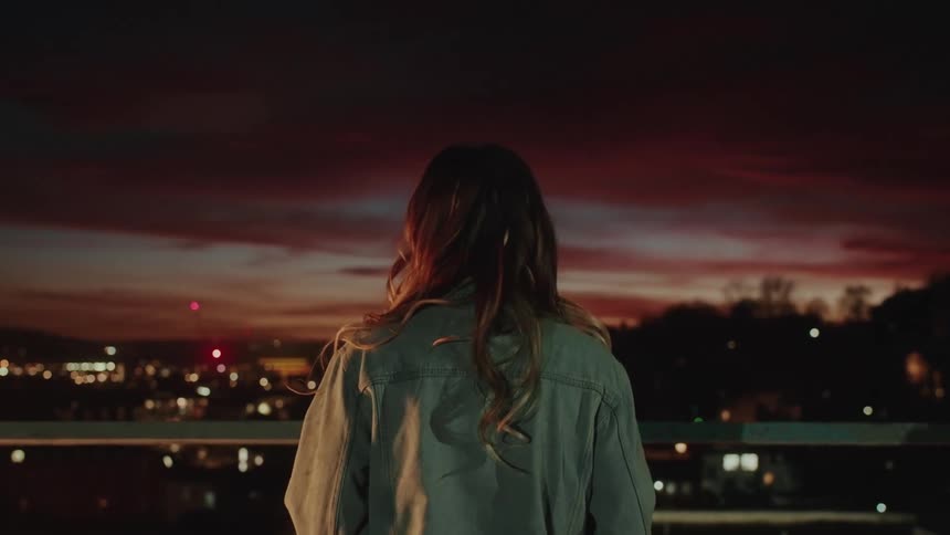Back view of a woman on a balcony overlooking a city at dusk