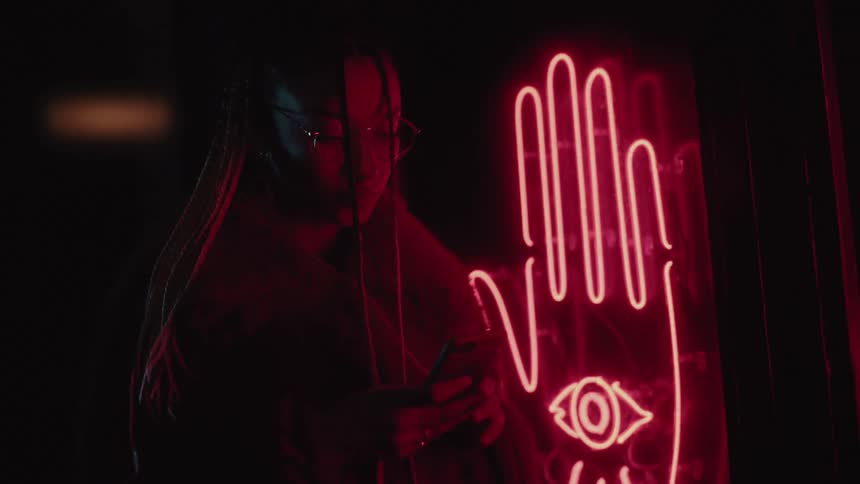 A woman checks her phone and walks past a neon sign of a hand