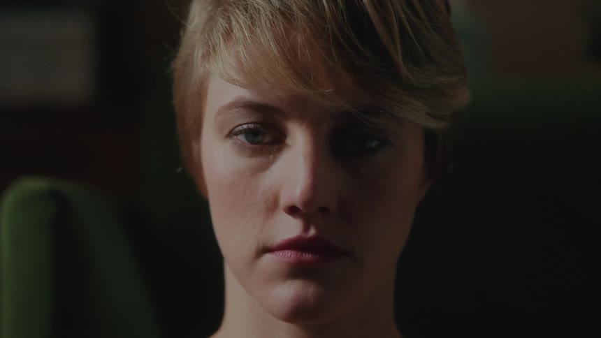 A dimly lit closeup of a young woman with green eyes and short blonde hair