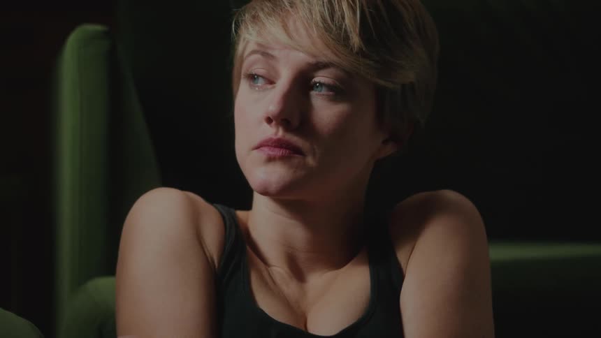 A blonde woman with short hair and blue eyes is looking to her right side while she caress her arms and she is sad