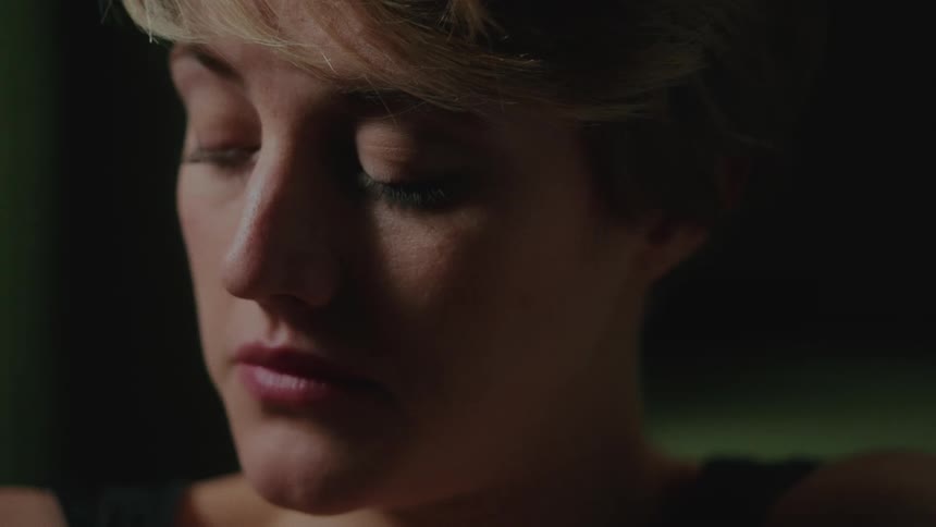 A dimly lit closeup of a young woman with green eyes and short blonde hair