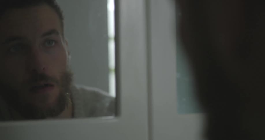 A young. bearded man raising his head to look at his reflection in the mirror and then rubbing his hand over his face