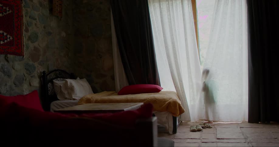 A child standing at an open window in a dimly lit bedroom with curtains billowing in the breeze and two beds visible in the background
