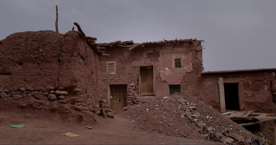 A dilapidated mudbrick house with a collapsed roof and crumbling walls surrounded by piles of rubble and overgrown vegetation