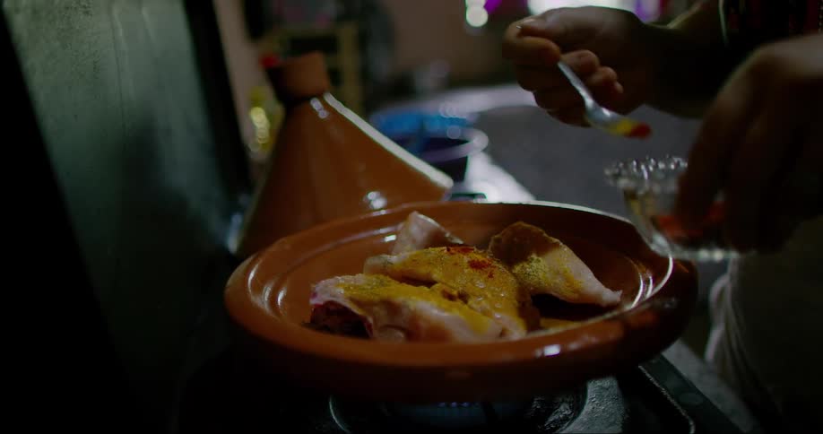 A hand is pouring chili powder over a dish in a tagine pot on a stove