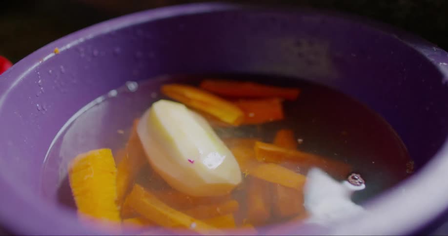 A hand placing a potato into a bowl of water filled with carrots