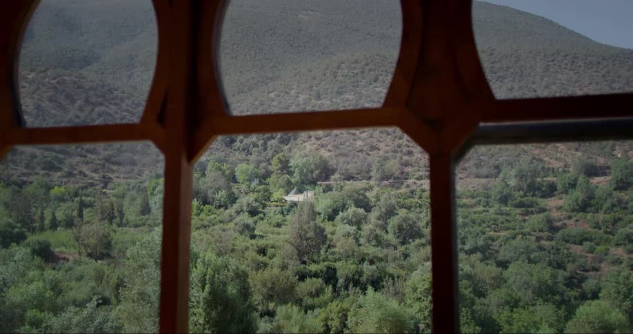 A view through a window with a wooden frame looking out onto a green landscape with dense vegetation and mountains