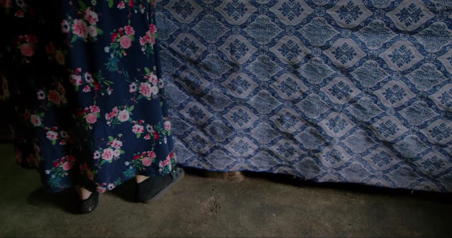 A woman in a house dress and slippers shimmies a bit while standing next to a bed